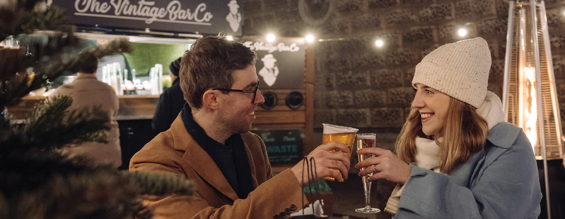 A young couple - man in a brown jacket holds a beer and woman in a blue jacket holds a prosecco